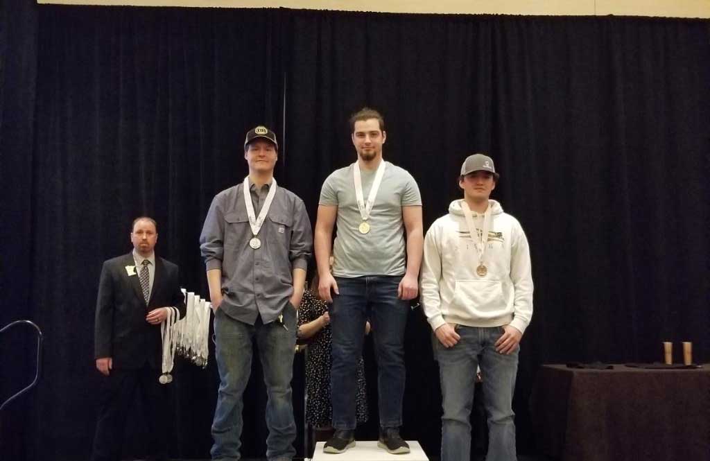 Three students standing on podium with man awarding medals in background