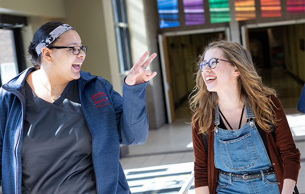 two female students walking through campus hallway talking and smiling