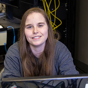 smiling student at laptop in IT classroom with network cables in background