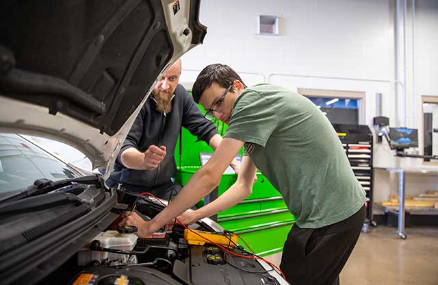 Student working under hood of car while faculty observes