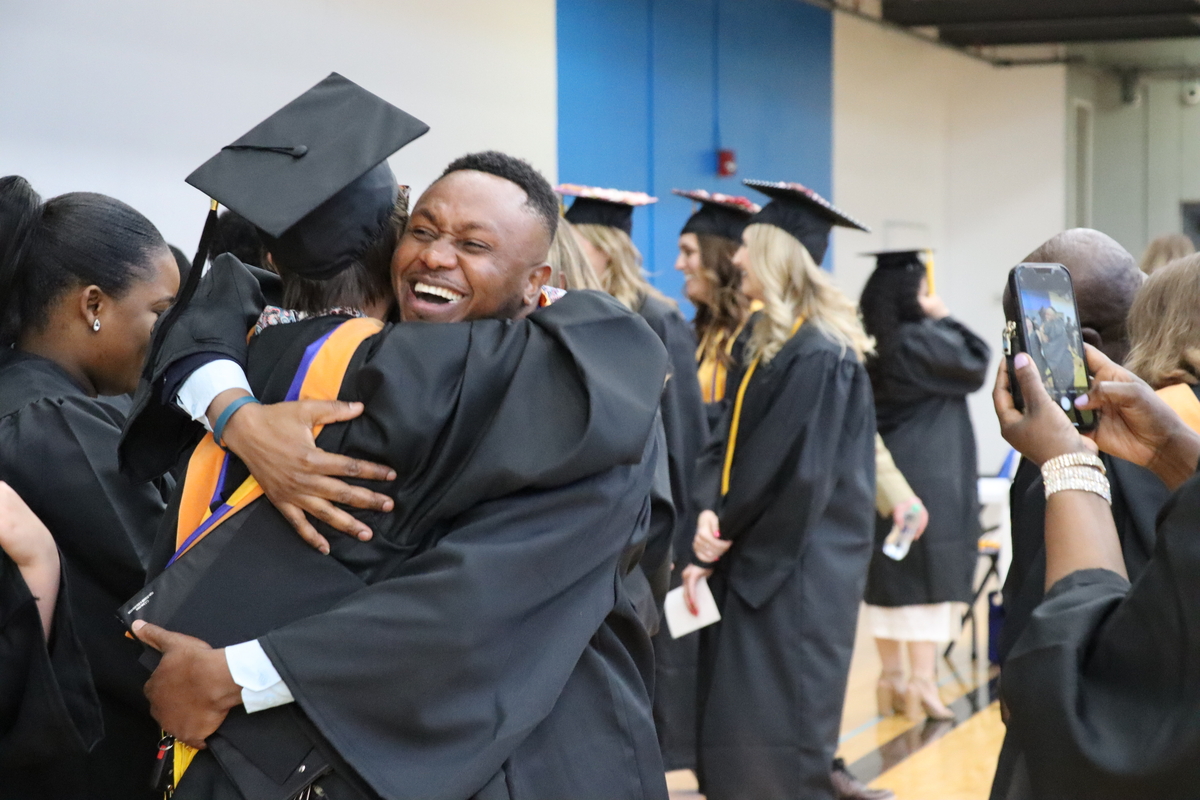 A joyful graduation scene with a graduate hugging another person amidst a crowd of fellow graduates and onlookers taking photos.