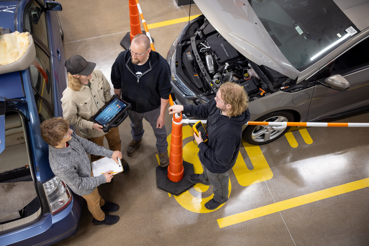 Four people discussing in a workshop with an electric car connected to diagnostic equipment. Two individuals are using digital tablets, all surrounded by safety cones.